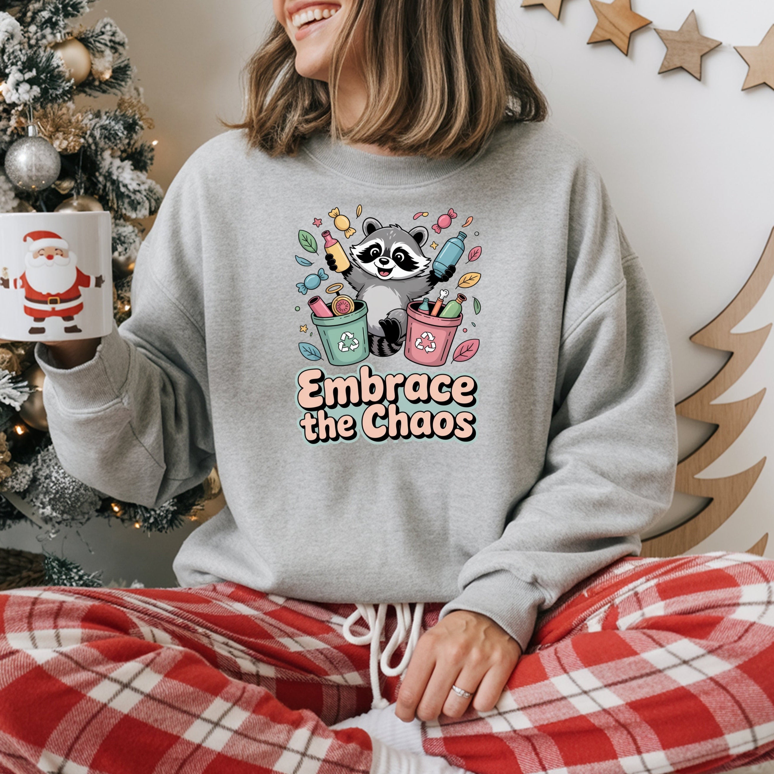 Person wearing a sweatshirt with a raccoon graphic and 'Embrace the Chaos' text, holding a mug in front of a decorated Christmas tree.