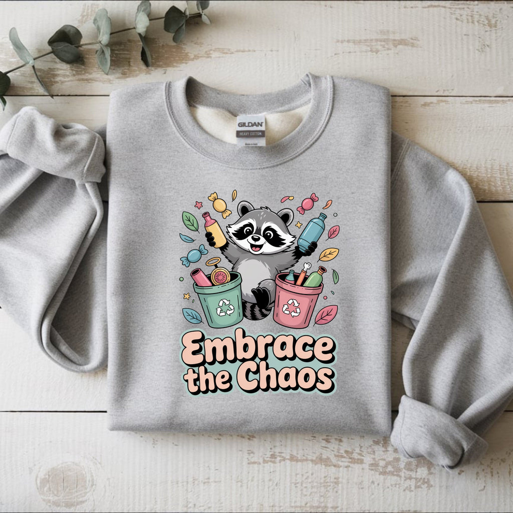 Gray sweatshirt with a raccoon graphic and 'Embrace the Chaos' text on a wooden surface.