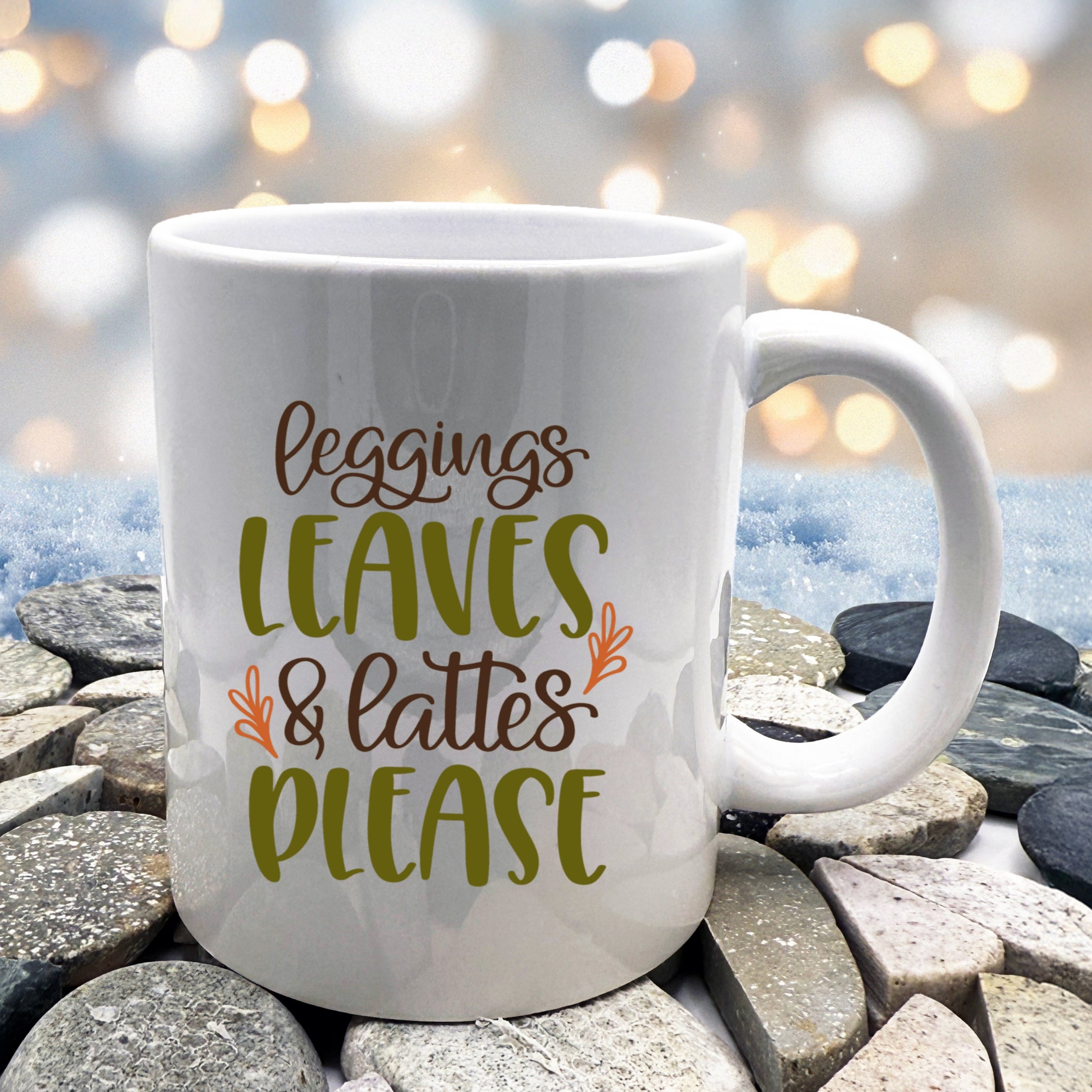 White mug with 'Leggings, Leaves & Lattes Please' text on a stone surface with blurred lights in the background