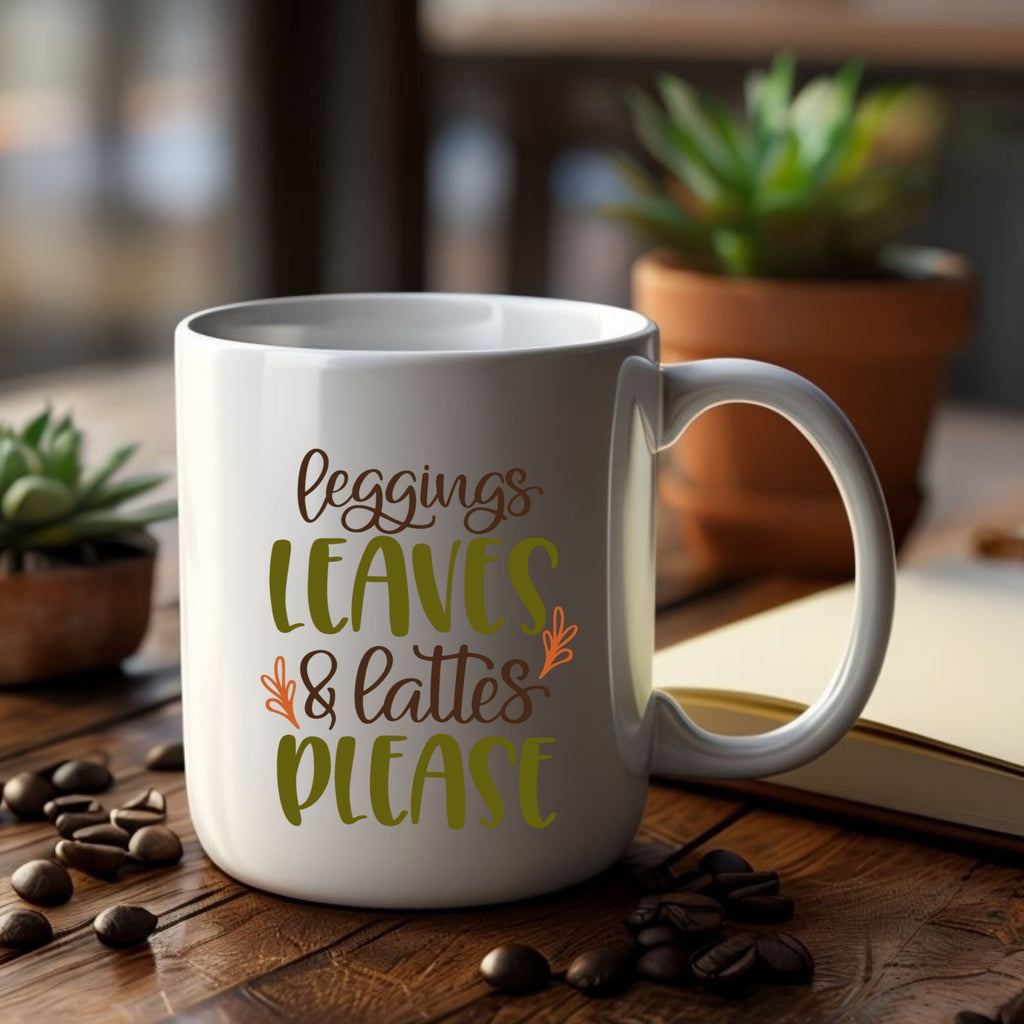 White mug with text 'leggings, leaves & lattes please' on a wooden table with coffee beans and plants.