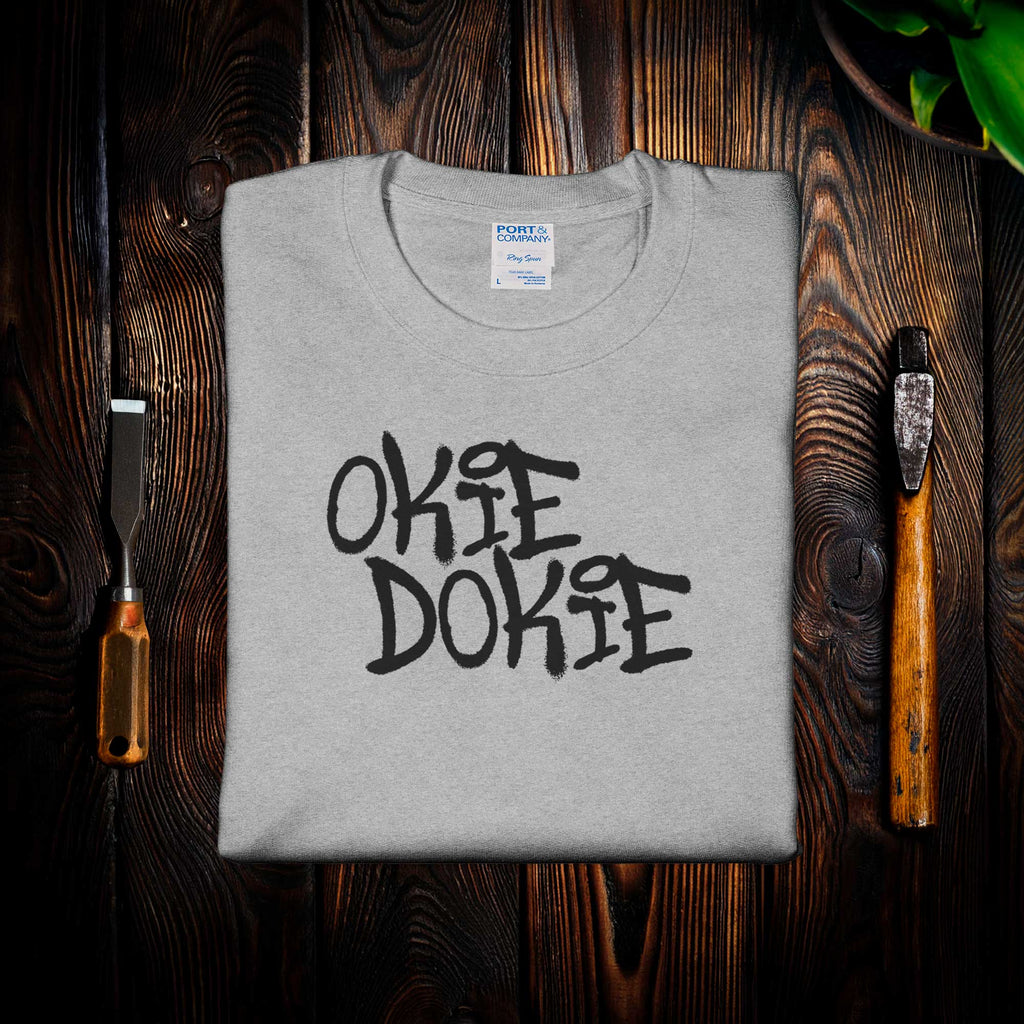 Fallout Amazon Show Gray t-shirt with 'OKIE DOKIE' text on a wooden surface with tools.