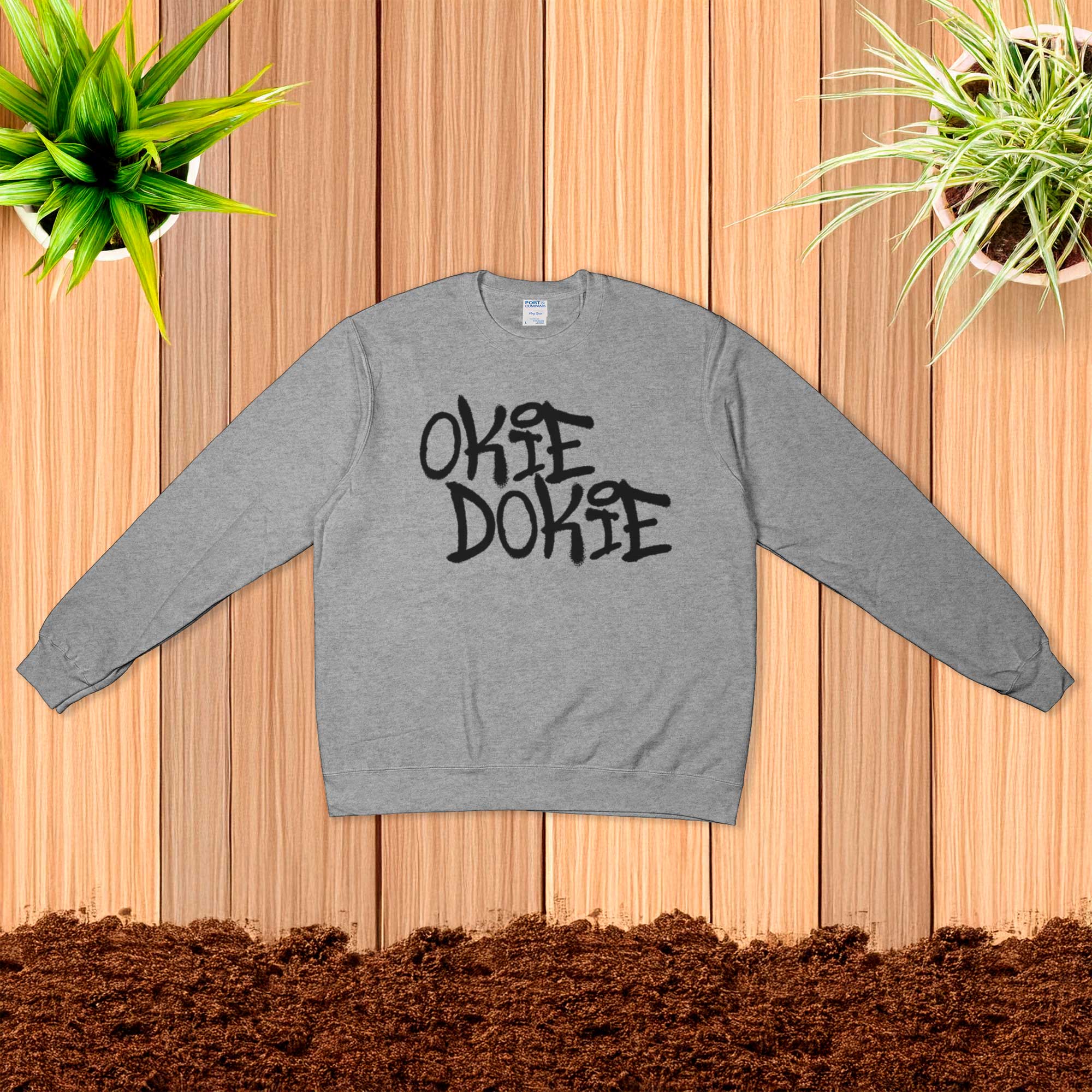 Fallout Amazon Show Gray sweatshirt with 'OKIE DOOKIE' text on a wooden surface with plants.