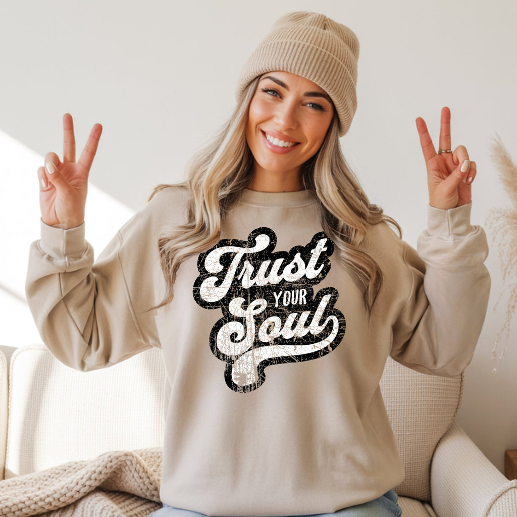 Smiling woman in beige Trust Your Soul unisex crewneck sweatshirt, beanie, making peace signs. Cozy casual wear.