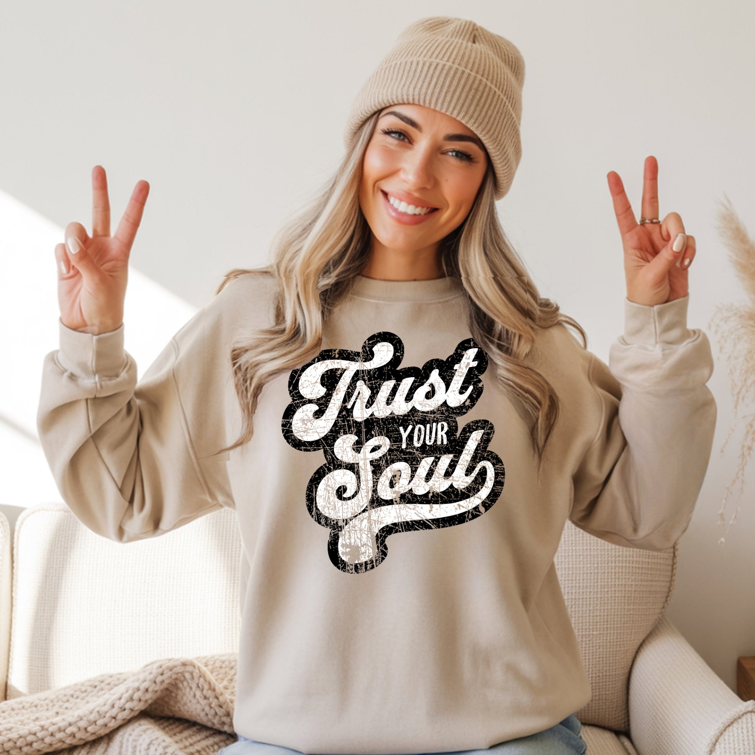 Smiling woman in beige Trust Your Soul unisex crewneck sweatshirt, beanie, making peace signs. Cozy casual wear.
