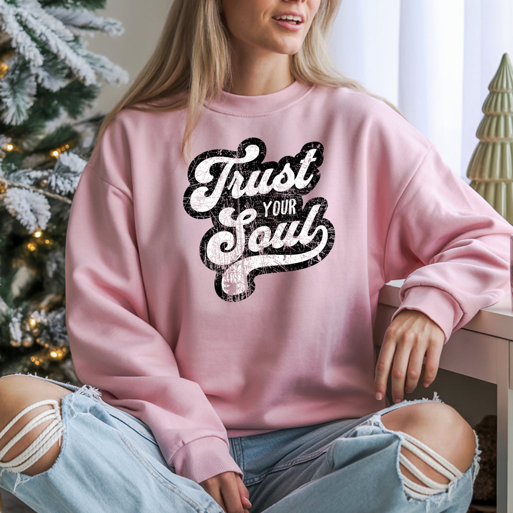 Person wearing a pink sweatshirt with 'Trust Your Soul' text in a festive setting with a Christmas tree.