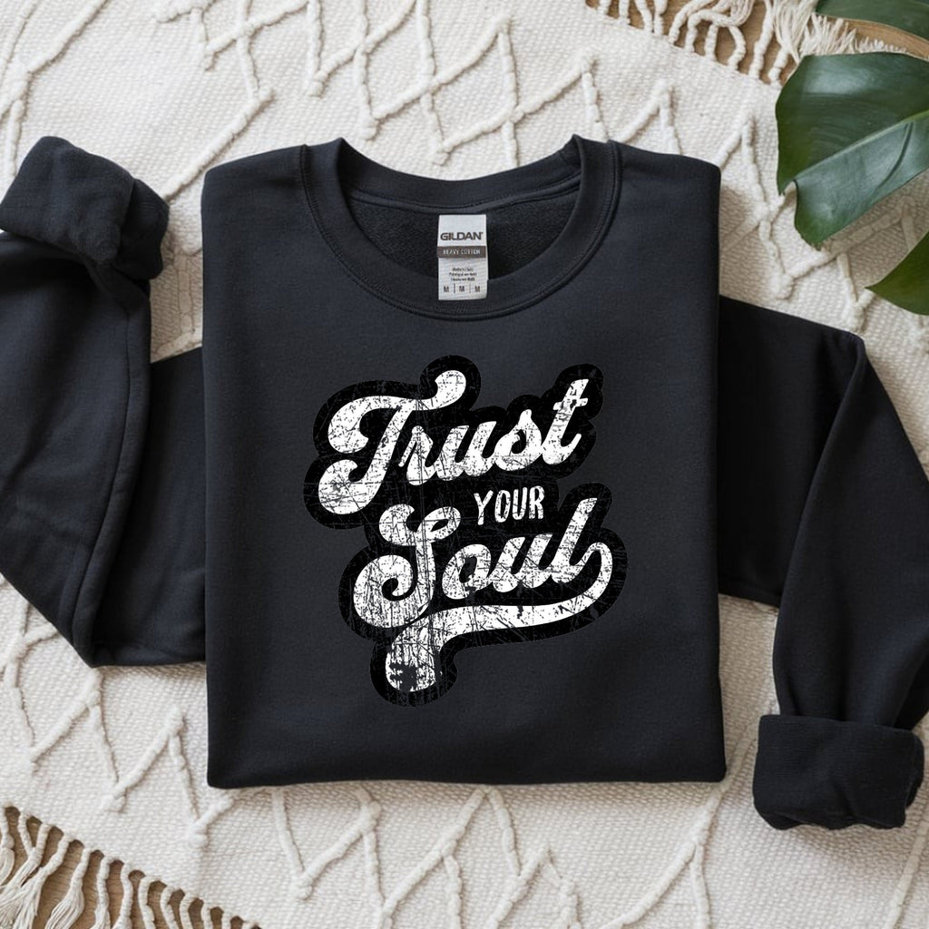 Black sweatshirt with 'Trust Your Soul' text on a textured surface with a leaf.