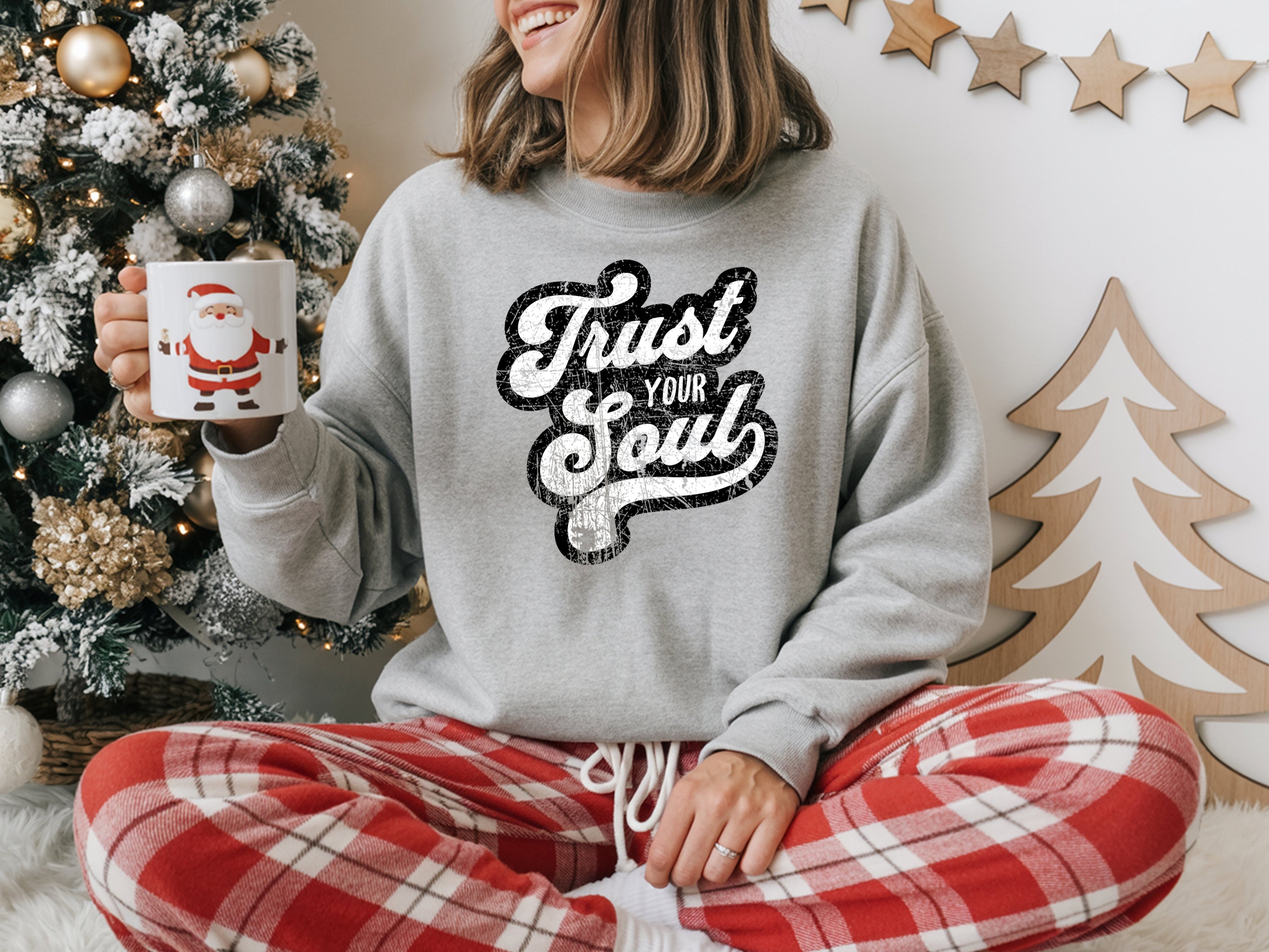 Person wearing a gray sweatshirt with 'Trust Your Soul' text and red plaid pants, holding a mug in front of a Christmas tree.