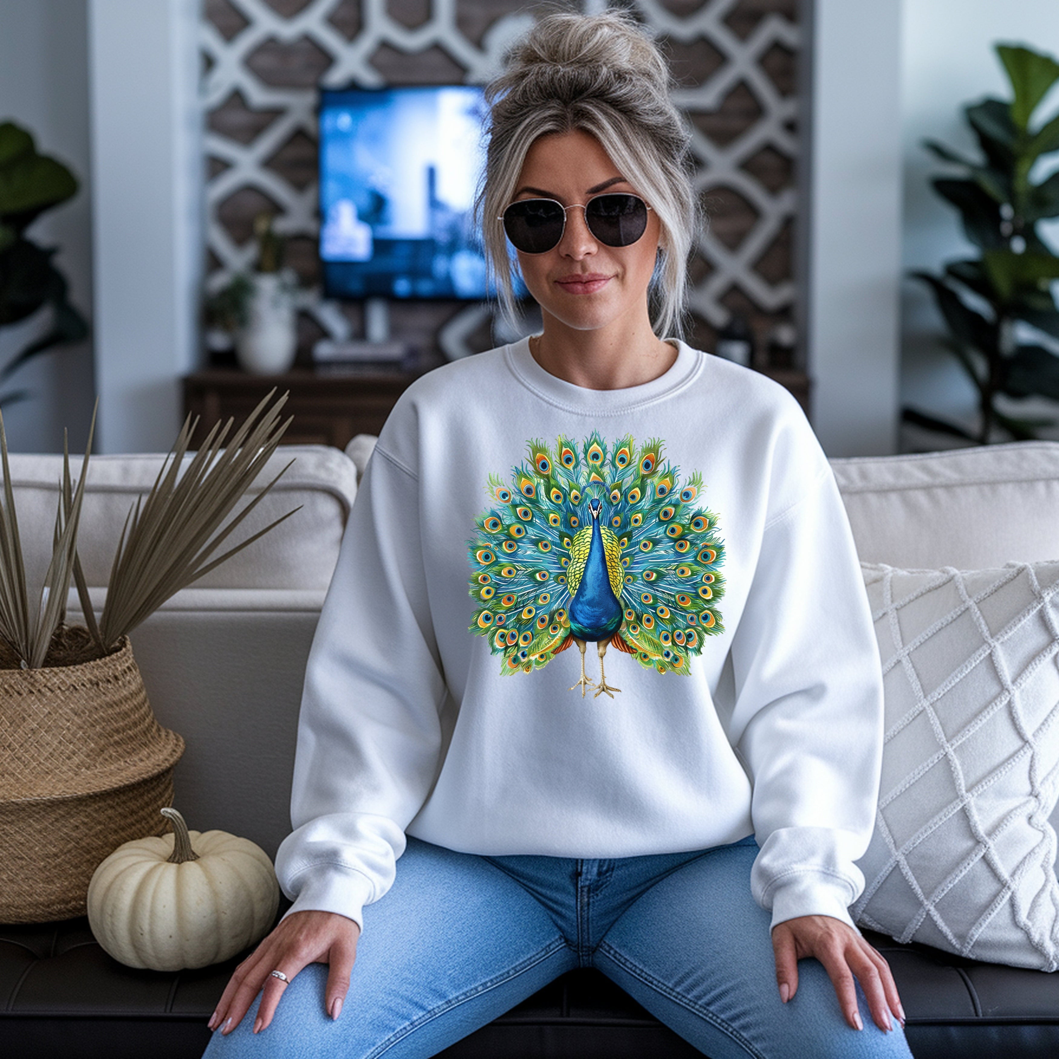 Woman wearing white unisex long sleeve tee with vibrant peacock design, blue jeans, and sunglasses, sitting on a couch.