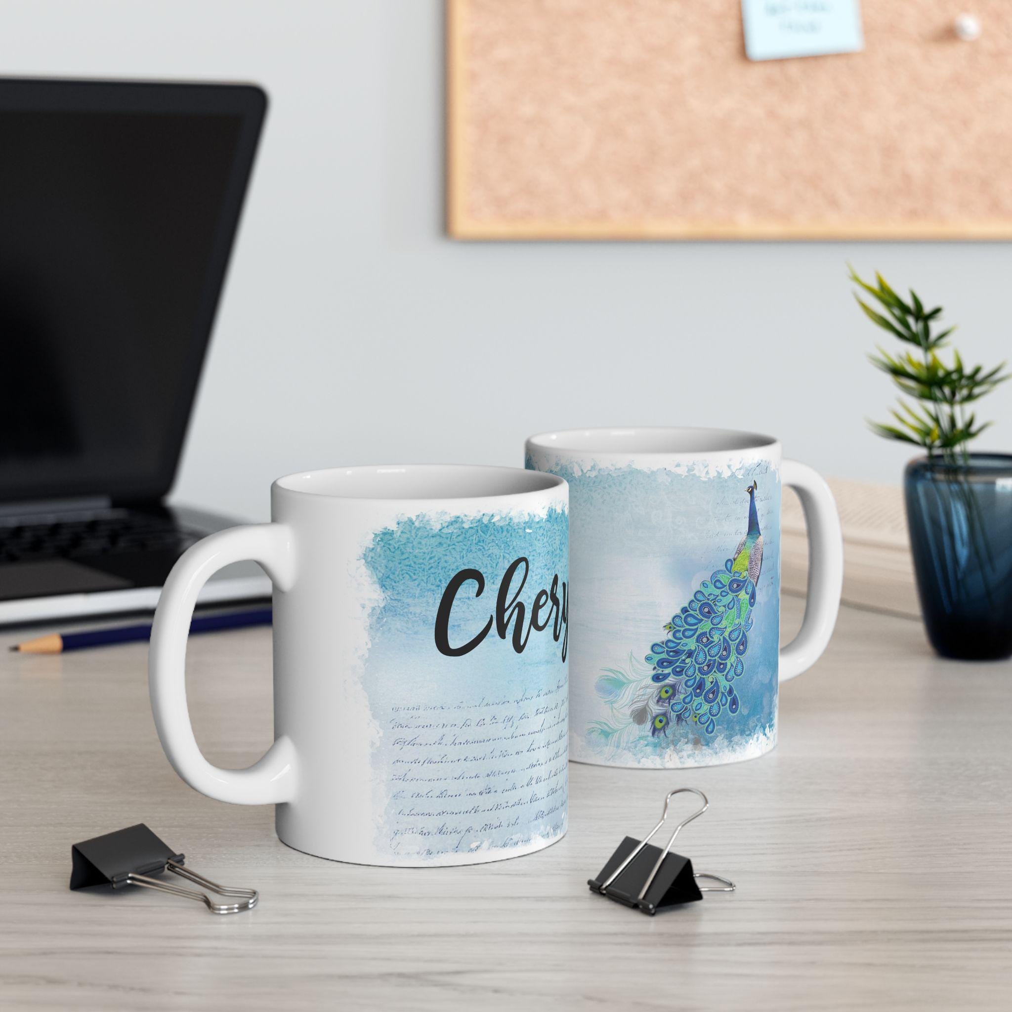 Two mugs on a desk with one featuring a peacock design and the other with text.