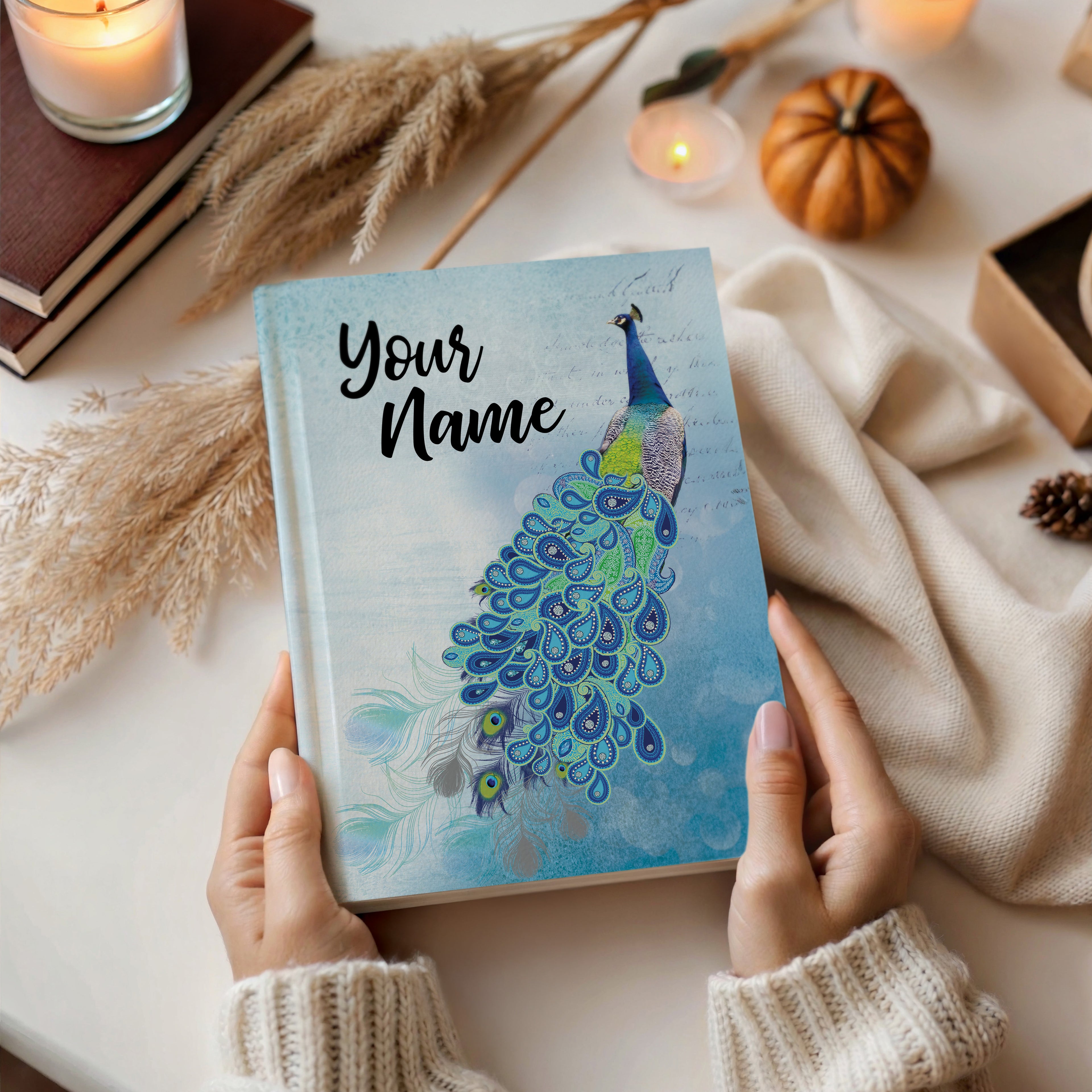Person holding a notebook with a peacock design and 'Your Name' text, surrounded by decorative items on a table.